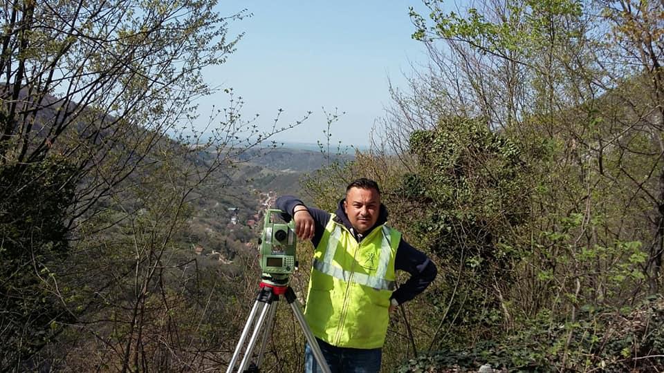 Membru al echipei PROMETER M&G lângă o stație totală, cu o vale împădurită în fundal